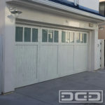 Pictured in this image is a double-car modern coastal cottage garage door in sandblasted white oak with a whitewash finish. The garage door features a one-to-three ratio with the top one-third consisting of three piece glass inserts of white-laminate glass panes. The bottom two-thirds of the garage door are vertical tongue and groove planks with a v-groove detail. The sandblasted, rustic surface of this door makes it resilient to its coastal environment and will improve in appearance as it ages. Blending the rustic oak material with the refined lines of the traditional craftsman style make this garage door a transitional piece of this coastal home's exterior that will be relevant for decades to come!