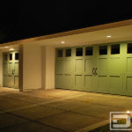 A double and a single car garage door in a shaker style. Each garage door has large, clear glass windows at the top panel. Vertical and horizontal trim pieces form a grid design with a decorative iron rivet at each intersection and a set of pull handles at the center to simulate an out-swing coach house door. The doors were finished in a bright pastel green color.