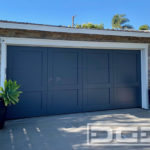 Pictured in this image is a double car garage door in a beautiful Shaker style. The garage door consists of four vertical panels with perimeter trim that divides the door in four equal parts. The top 2/3's of each panel is a recessed panel that is weighed in at the bottom with a trim piece running horizontally about 1/3 of the way up. The garage door is finished in a dark blue-gray color.