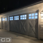 A double-car and a single-car garage door set in a traditional Craftsman farmhouse style. The garage doors are divided into vertical panels with a set of six glass pieces at the top that are divided with true mullions. The bottom 2/3's of each vertical panel boast a cross-buck design. Both doors were beautifully finished in a dark charcoal color and the windows were fitted with clear glass panes.