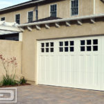 A coastal cottage double-car garage door in pure white. The garage door features a four vertical panel design with a cluster of six glass panes at the top individually divided by true mullions and fitted with clear glass. The bottom two-thirds of each panel consists of three vertical, narrow recessed panels while the rest of the trim work on the perimeter of each panel is framed with a six inch trim. This garage door design is perfect for a craftsman, shaker or transitional style home!