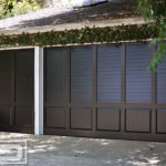 A double and a single car garage door set in a plantation shutter style. The garage doors feature a louvered top with perimeter trim work while the bottom of each door is weighed in with raised panels. The doors are finished in a satin black color that adds richness and interest to the garage doors. Each of the garage door openings is beautiful surrounded with mature vines that frame these louvered garage doors quite nicely!