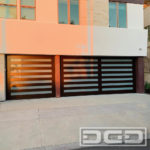 A double and a single car garage door project in a Contemporary style. The doors feature a horizontal open plank design fitted with white-laminate glass between the gaps. The garage doors are built out of composite engineered materials with a faux heart-grain finish that was applied by hand.