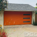 Pictured in this photo is a double-car Mid Century modern garage door in an orange color. The garage door features a horizontal, narrow plank design with a v-groove detail between each piece. On the right of the door are three vertical, narrow windows stacked on top of each other. Each window is fitted with a dark tinted glass pane for privacy and aesthetic purposes. This custom garage door can be finished in any color, the windows can be placed in a different configuration and the glass panes can be any designer type glass available in the market. This is the perfect garage door design for a Mid Century Modern or Contemporary style home!