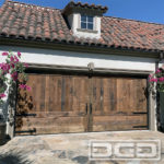In this image, there is a double-car garage door in authentic reclaimed wood. The sectional garage door boasts a carriage door style by configuring the trim work to resemble two large out-swing leaves that would part in the middle. The top one-third of the door is composed of vertical tongue and groove planks while the bottom one-third boasts two large pyramid panels, one on each side, that finish off the garage door’s design. Exquisite hand-forged decorative hardware includes two pull handles at the center trim pieces to accentuate the simulated carriage door partition. Six decorative strap hinges, three on each end of the door finalize the old-world carriage door look of this custom reclaimed wood garage door. The reclaimed wood that was used to build this Vintage Nouveau 03 garage door adds plenty of old-world character and will certainly capture the attention of passers-by for years to come!