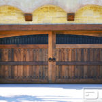 This is an image of a double-car Tuscan architectural garage door crafted in real reclaimed vertical grain Douglas fir lumber. The overhead garage door is trimmed out to simulate two large out-swing carriage door leaves. The top one-third of the door boasts an arched dual-pane window that spans the entire width of the garage door. Each of the two windows features an iron-forged grill insert and they are both fitted with designer antique seedy glass panes that tie in with the rustic nature of the reclaimed lumber used to handcraft this garage door. At the center of the door, there is a set of ring pulls that emphasize the carriage door partition. Several rows of diamond-shaped clavos complete the Tuscan style of the garage door. The reddish tonalities seen throughout the door’s surface are due to the reclaimed Douglas fir lumber species that was used to manufacture the door itself. When closed, this garage door really fools the eye by appearing to be an out-swing carriage door but it is a stunning surprise to see it roll up in sections like a standard sectional garage door!