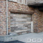 Photographed in this image is a modern industrial-style garage door that is handcrafted in real reclaimed barn wood. The door features a staggered, horizontal plank design that is composed of two different reclaimed wood species. The silver/gray boards are reclaimed fir while the darker boards are reclaimed oak. The boards were all alternated throughout the garage door’s surface to create an abstract appearance that is consistent throughout the overhead garage door’s surface. Although the garage door design is simple, it is a complex creation that required careful planning and skilled craftsmanship to achieve!